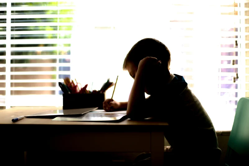 A boy sitting alone at a desk by a sunny window, his head resting on one hand and a pencil on his homework.